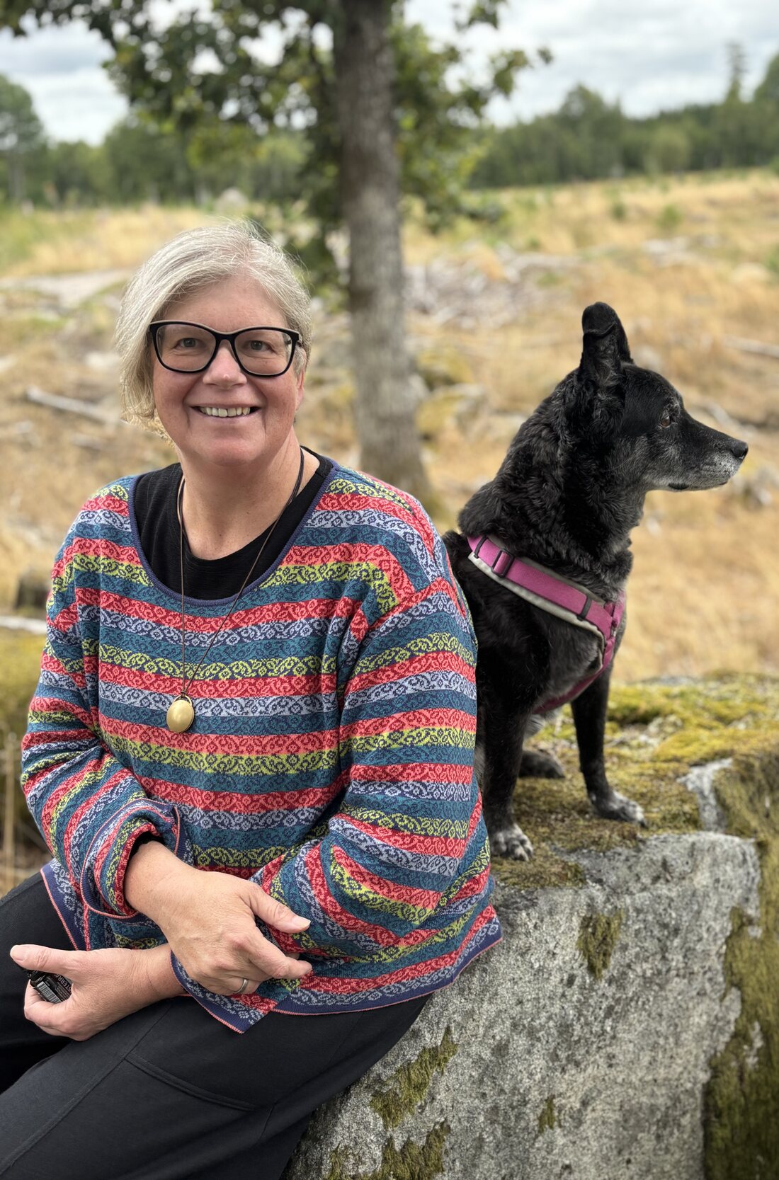 Nicola sitzt lächelnd neben ihrer Hündin Lucy auf einem großen Stein in der Natur